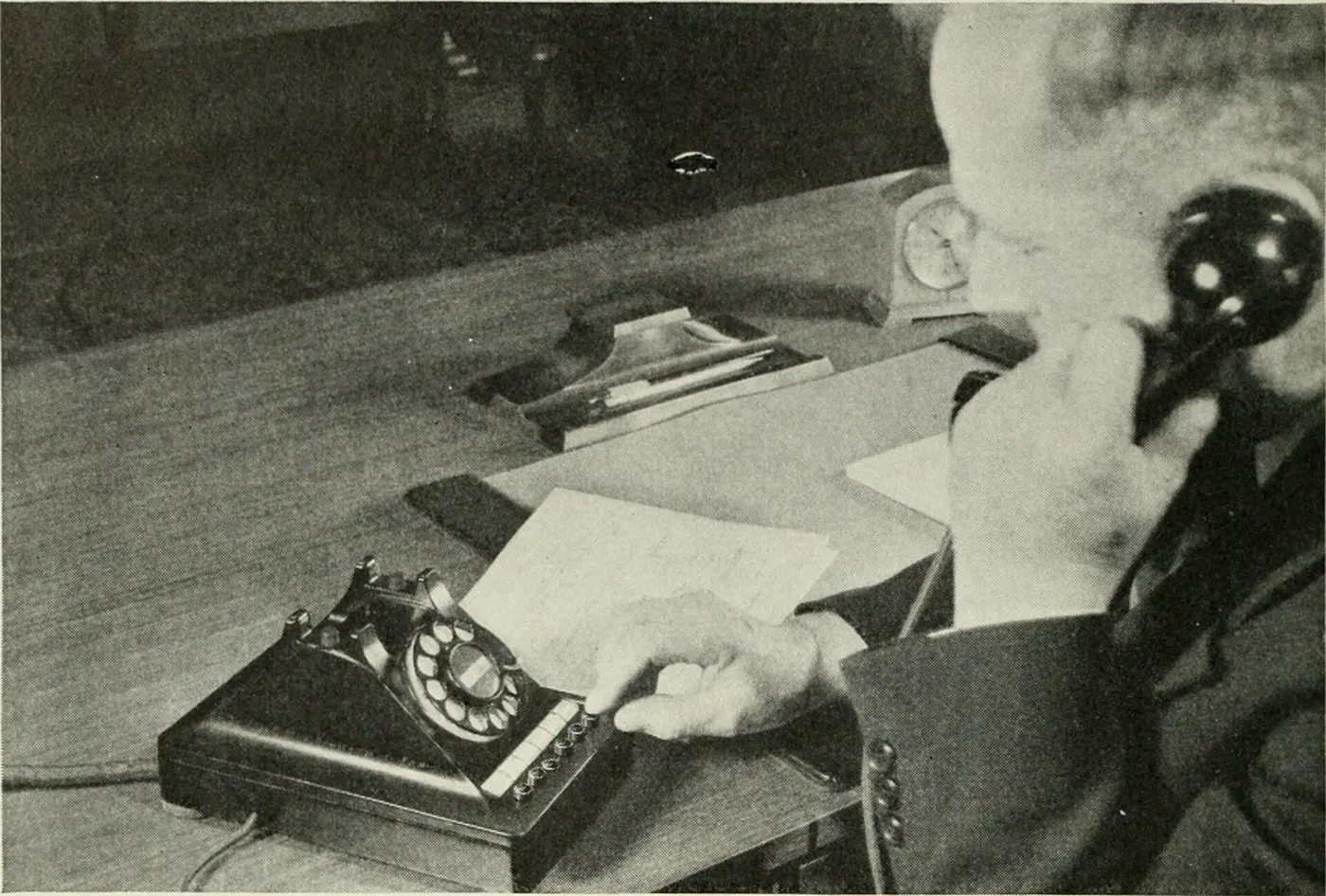 A man at a desk dials a rotary telephone while holding the receiver, from a 1940 Bell Telephone Magazine